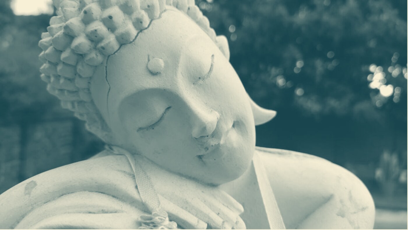 A cream stone Buddha statue, eyes closed, resting its head on its hand, in a garden at the centre