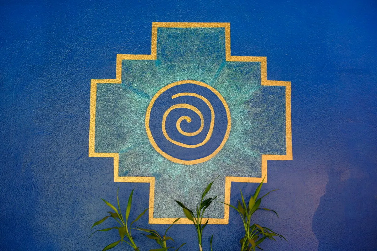 A gold and turquoise spiral set in a stepped cross, painted on a deep blue wall above young bamboo shoots